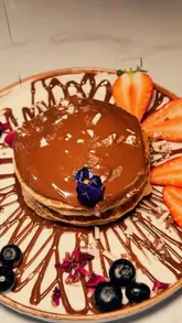 A visually appealing montage showcasing the food, drinks, and floral-themed interior of La Dolce London. The video features close-up shots of Eggs Benedict, chocolate-drenched pancakes, cannoli, and coffee art, interspersed with wide shots of the cafe's aesthetic decor, including chandeliers and floral ceiling displays.