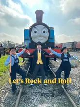 A bright, well-composed promotional shot featuring three costumed characters standing on railway tracks in front of a Thomas the Tank Engine steam train. The image effectively captures the theme of the attraction, using a symmetrical composition that highlights the venue's main draw.