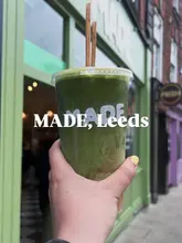 A first-person perspective shot holding a plastic takeaway cup containing a layered drink (half matcha, half iced coffee) in front of a green-fronted cafe in Leeds.