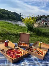 A high-quality, bright, and inviting outdoor shot of a picnic featuring takeaway pizza boxes from Pizzarova, set against the iconic backdrop of the Clifton Suspension Bridge in Bristol. The composition is well-balanced, using the natural light to highlight the food while providing a strong sense of place.
