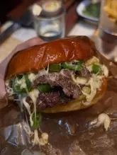 Here are a few options for your alt text, keeping it descriptive and SEO-friendly:

**Option 1 (Descriptive):**
Close-up of a juicy Flat Iron green chilli burger served on a wooden board with a side of fries in a London restaurant.

**Option 2 (Action-oriented):**
A delicious green chilli burger from Flat Iron steakhouse in London, served with a side of crispy golden fries.

**Option 3 (Concise):**
Flat Iron London signature green chilli burger with fries on a wooden serving board.

**SEO Tip:** Alt text is best when it describes what is actually in the image for visually impaired users while naturally incorporating your main keywords (Flat Iron, burger, London).