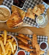 An overhead, flat-lay photograph of a pub-style sharing spread featuring various fried items, bread, and dips served on wooden boards with checkered paper.