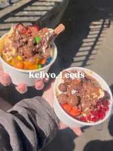 A high-angle, first-person perspective shot of two cups of frozen yogurt held by two people, showcasing a variety of toppings including fresh fruit, chocolate sauce, and candy. The image is captured outdoors in bright, direct sunlight.