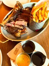 A top-down, overhead shot of a Côte de Boeuf sharing platter served with a side of fries and three different sauces, presented on a wooden table.