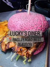 Here are a few options for your SEO alt text, depending on which aspect of the image you want to highlight:

**Option 1 (Focus on the burger):**
Double meat Lucky’s Grilled Stack burger with a pink bun and peri fries at Lucky’s Diner on Paisley Road West, Glasgow.

**Option 2 (Focus on the full experience):**
A delicious spread of burgers, fries, and a massive chocolate Matilda cake at Lucky’s Diner in Glasgow.

**Option 3 (SEO-focused):**
A mouthwatering burger meal and decadent chocolate cake at Lucky’s Diner, a popular restaurant in Glasgow.

**Best Practice Tip:** If the image shows a specific person or a clear view of the food, use the one that most accurately describes what the viewer sees first!
