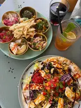 An overhead, natural-light shot of a lunch spread featuring a mezze platter, a halloumi flatbread, and iced tea, set on an outdoor table.