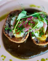 A close-up, top-down shot of a scotch egg served on a dark green sauce, garnished with pickled red onions and microgreens, presented on a pale pink plate.