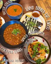 A flat-lay shot of a variety of Indian-inspired small plates on a wooden table, featuring dishes like butter chicken, lamb birria-ish tacos, and spiced cauliflower.