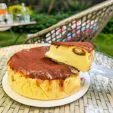 A close-up, high-angle shot of a caramel tiramisu Basque cheesecake being lifted with a cake server, set on a glass table in an outdoor garden setting.