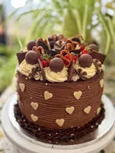 A high-quality, close-up photograph of a decadent, multi-layered chocolate cake decorated with various toppings including chocolate truffles, pretzels, strawberries, and white chocolate hearts. The cake is presented on a white cake stand, with a soft-focus background.