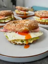 A close-up, appetizing shot of a breakfast bagel featuring a runny egg yolk, avocado, ham, and cheese. The image is well-lit with natural light and uses a shallow depth of field to focus on the texture of the bagel and the dripping yolk.