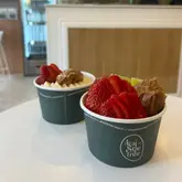 A close-up shot of two acai bowls served in branded paper cups, featuring toppings like strawberries, banana, kiwi, and nut butters. The bowls are placed on a white table in a brightly lit, minimalist cafe setting.