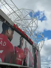 A clear, daylight shot of the exterior of Old Trafford stadium, featuring a large promotional banner of Manchester United players against a blue sky.