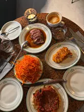 A high-quality, overhead flat-lay shot of a table spread at a restaurant, capturing a variety of dishes including lamb, a carrot salad, a tomato flatbread, and a fried spam dish. The lighting is warm and moody, effectively conveying the intimate, vibey atmosphere of the venue.