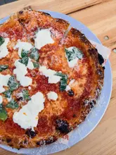 A top-down, natural-light shot of a Margherita pizza topped with stracciatella, served on a metal tray on a wooden table. The image captures the texture of the crust and the fresh ingredients, though the framing is quite tight.