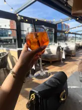 A bright, inviting POV shot of an Aperol Spritz held up against a sunlit, outdoor terrace setting, effectively capturing a summer atmosphere.