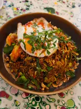 A top-down, overhead shot of a Nasi Goreng dish served in a dark bowl, topped with a fried egg and garnished with fresh herbs and fried shallots. The dish is presented on a patterned table surface.