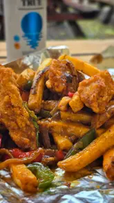 A short, handheld video showcasing a 'spice bag' takeaway meal from a food truck. The creator opens the branded paper bag to reveal the contents, including chicken strips, seasoned fries, peppers, onions, and a side of curry sauce, paired with a non-alcoholic lager.