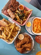 A top-down flat lay photograph of a spread of casual comfort food, including fried chicken, fries, mac and cheese, and a hoagie, served on a blue table.