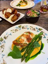 A high-quality, well-composed food photograph featuring a beautifully plated monkfish dish with asparagus and a vibrant sauce, accompanied by side dishes on a rustic wooden table. The lighting is soft and natural, highlighting the textures and colours of the food, creating an inviting and sophisticated dining atmosphere.