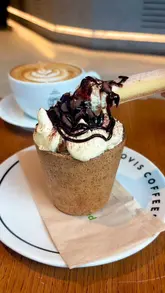 A short, aesthetic video showcasing a 'Cookie Cloud Cup' and a latte at Ovis Coffees. The video features close-up shots of the cookie cup being topped with cream and chocolate sauce, followed by a latte with latte art, and a spoon interacting with the dessert.