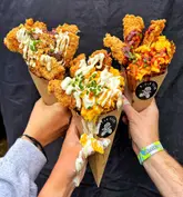 A close-up, first-person perspective shot of three people holding street food cones filled with fried chicken and mac and cheese, set against a plain black backdrop.