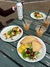 A casual, overhead shot of a brunch spread on a weathered outdoor table, featuring avocado toast with halloumi, a toasted sandwich with chips and salad, and iced coffees.