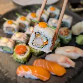 A close-up, first-person perspective shot of a piece of uramaki sushi being held with chopsticks, with a larger platter of various sushi pieces blurred in the background. The image highlights the freshness of the sushi, specifically the shrimp tempura roll being held, and provides a clear view of the variety available in the bottomless brunch offering.