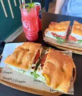 A casual, well-lit photograph of two focaccia sandwiches and a strawberry lemonade served on branded wooden boards at a cafe.
