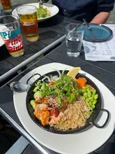 A casual, outdoor shot of a teriyaki salmon poke bowl served in a metal pan, accompanied by a beer and water glass on a black table.