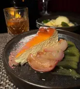 A high-quality, moody food photograph featuring a slice of basque cheesecake with poached fruit, served in a restaurant setting.