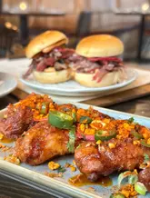 A high-quality, close-up food shot featuring a plate of glazed chicken wings topped with chili and spices in the foreground, with a pair of sliders in the blurred background. The image uses a shallow depth of field to draw focus to the texture and glaze of the wings, creating an appealing and appetizing visual.