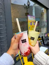 A first-person perspective shot holding two vibrant bubble tea drinks in front of a shop window, showcasing the product clearly.