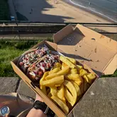 A first-person perspective shot of a takeaway fish and chip meal, featuring cod tacos and chips, held against a scenic coastal backdrop.