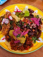 A top-down, overhead shot of a trio of vegan tacos served on a bright yellow plate. The tacos are generously topped with various ingredients including pickled onions, radish, and fresh herbs, set against a simple wooden table background.