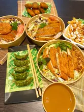 A flat-lay photograph of a takeaway spread featuring various Japanese-inspired dishes including katsu bowls, gyoza, and fries, presented in disposable containers on a bamboo mat.