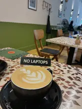 A close-up, eye-level shot of a latte with latte art served in a black cup on a mosaic-tiled table. A prominent 'NO LAPTOPS' sign is placed directly behind the coffee, dominating the center of the frame.