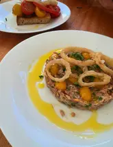 A top-down shot of a steak tartare dish served with fried shallot rings, accompanied by a partially visible bruschetta on a separate plate in the background, set on a wooden table.