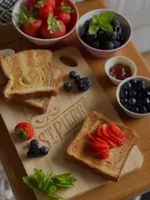 A bright, professionally styled overhead shot of a breakfast spread featuring toasted croissant loaf topped with fresh strawberries, accompanied by bowls of fresh berries. The image uses natural light to highlight the textures of the food, creating an inviting and appetizing home-cooking scene.