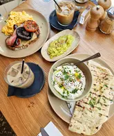 A bright, overhead shot of a brunch spread featuring a plate of scrambled eggs with bacon, sausage, mushroom, and tomato, alongside a bowl of poached eggs with yoghurt and flatbread, and iced coffees.