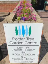 A clear, well-lit photograph of a sign for Poplar Tree Garden Centre, set against a backdrop of flowers. The image provides essential information about the venue, including opening hours, and establishes the location's identity.