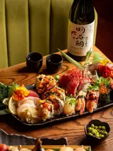 A high-quality, professional food photography shot featuring a large, beautifully arranged sushi and sashimi platter, accompanied by a bottle of Akashi-Tai sake and a small bowl of edamame. The lighting is warm and inviting, highlighting the textures and colours of the fresh fish and garnishes, set against a dark wooden table and a green upholstered booth.