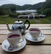 A serene, eye-level shot of a teapot and two cups of herbal tea on a wooden table, set against a picturesque backdrop of a lake, boats, and rolling hills in the Lake District.