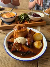 A high-quality, appetizing close-up shot of a Sunday roast dinner, featuring a generous portion of beef brisket, a large Yorkshire pudding, and roast potatoes. The image captures the rustic, hearty nature of the meal, though the background is slightly cluttered with other diners and table items.
