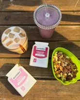 A lifestyle flat-lay featuring a morning routine with an iced latte, a berry protein shake, a bowl of mixed nuts, and two supplement bottles on a wooden table.