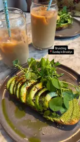 A short, handheld-style reel showcasing a brunch visit to 'Sunday in Brooklyn'. The video features close-ups of avocado toast topped with halloumi and iced lattes, followed by brief shots of the restaurant's interior bar area and the exterior facade.