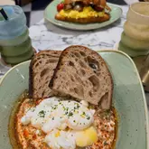 A top-down, overhead shot of a brunch table featuring a plate of poached eggs on a spiced yogurt base with sourdough toast, accompanied by matcha lattes and a pastry in the background.
