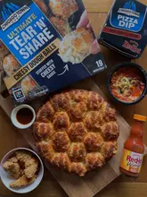 A clean, overhead product shot featuring a Chicago Town Tear 'N' Share pizza, a Frank's RedHot Buffalo Chicken Pizza Dip, and a bowl of chicken tenders arranged on a wooden board.