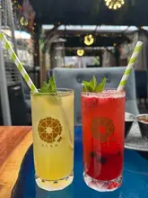 A bright, eye-level shot of two colourful mocktails in branded glassware, set against a blurred outdoor courtyard background at Alam restaurant.