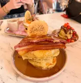 A close-up, eye-level shot of a stack of pancakes topped with bacon, served on a marble table in a cafe setting. The image captures a casual dining moment with other plates and personal items visible in the background.